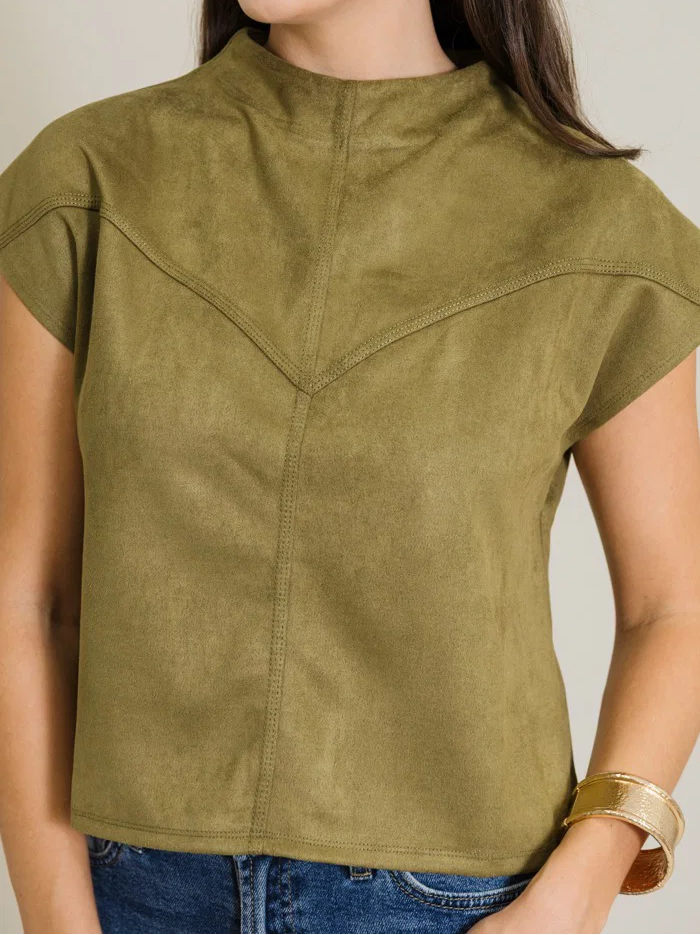 A woman wearing a cropped short-sleeve top in olive green faux suede, paired with blue jeans and gold jewelry, against a neutral beige background.