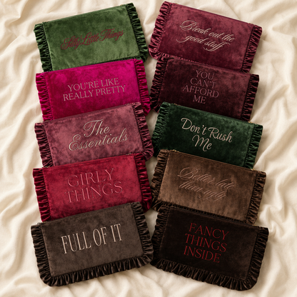 A set of rectangular velvet cosmetic bags with ruffle edges, arranged in a grid on a cream satin fabric background. Each cosmetic bag features a different color (green, burgundy, magenta, mauve, brown, and charcoal) with matching embroidered text on the front.